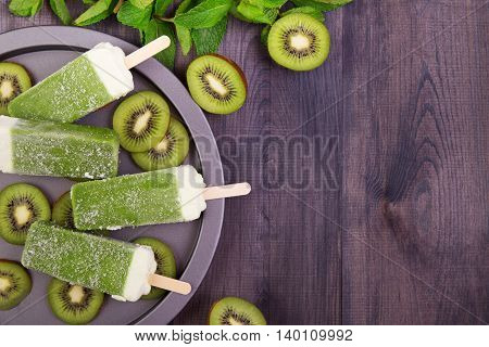 Homemade frosty kiwi popsicles on a plate with mint and kiwi slices on a dark wooden table. Top view with copyspace