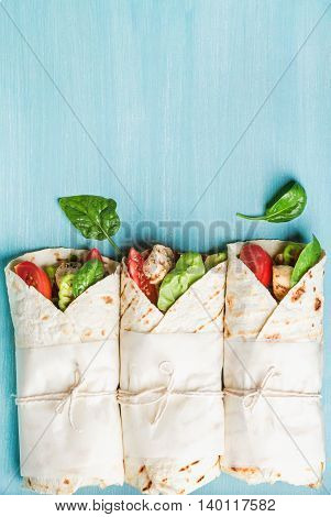 Healthy lunch snack. Three tortilla wraps with grilled chicken fillet and fresh vegetables over turquoise blue painted wooden background. Top view, copy space, vertical composition