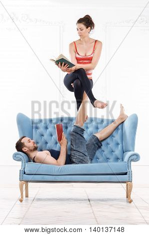 Young Beautiful Man And Woman Practicing Acroyoga On Blue Sofa Doing Excercise And Reading Books In White Interior