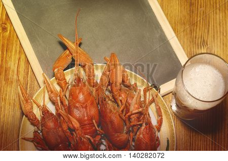 Boiled crawfish, glass of light beer and black chalkboard on a wooden table. Top view, vintage filtered picture.