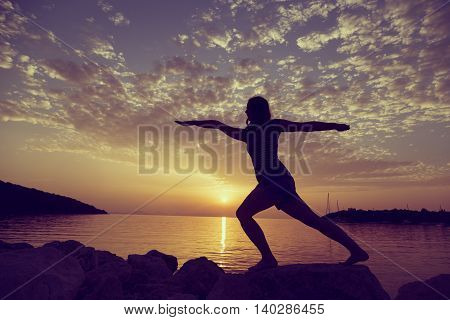 Silhouette of a young woman practicing yoga on a rocky shore in Syvota bay Greece. Cloudy sky Ionian sea and a beautiful sunset in the background.