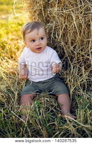 gay boy kid blonde in white tank top sitting on a field of hay next to the stack during sunset