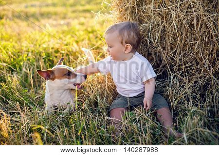 gay boy kid blonde in white tank top sitting on a field of hay next to the stack with the dog during sunset