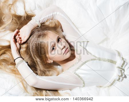 small girl kid with long blonde hair and pretty serious face in prom dress lying on white fabric closeup