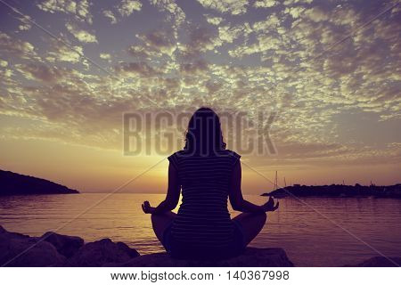 Silhouette of a young woman practicing yoga on a rocky shore in Syvota bay Greece. Cloudy sky Ionian sea and a beautiful sunset in the background.