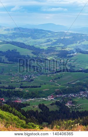 landscape consisting of a Carpathians mountains with fir-tree and green grassy valley with houses and blue sky with white clouds on the background