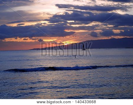 Dramatic Sunset dropping behind the Waianae Mountain range with clouds waves and boats on the Pacific ocean off the coast of Oahu Hawaii.