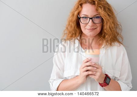 Warm up. Cheerful beautiful senior woman holding thermos with tea and expressing joy while standing isolated on grey background