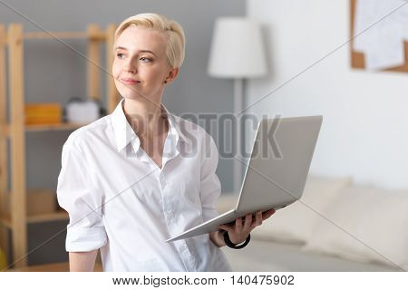 Have a look. Cheerful confident worker standing and holding a laptop while looking aside and smiling