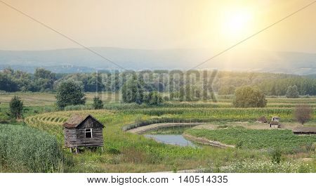 Rural landscape of old log cabin beside small river and fields in plains