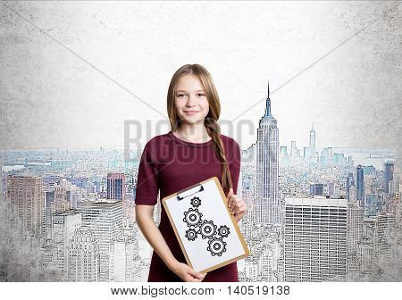 Young lady is standing in red dress with braided hair and holding clipboard with gears sketch on it. Background is painting of New York. Concept of strategy importance in business