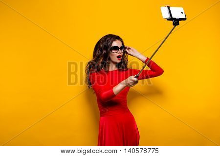 Portrait of beautiful brunette in awesome dress and red lips wearing sunglasses while making selfie with stick. Isolated