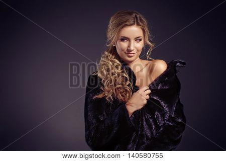 Fashionable young woman with long blonde hair smiling at camera while wearing black fur coat. Studio shot.Isolated.