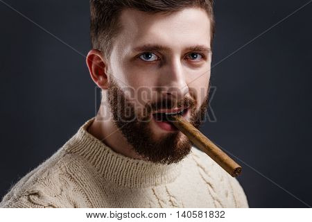 Portrait of handsome serious bearded man in white sweater holding cigar on black background