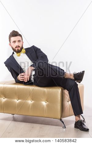 Portrait of handsome man relaxing on modern pouffe in elegant suit with bow-tie