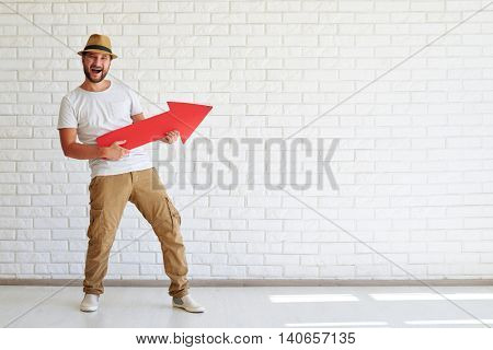 Happy keenness stylish man are standing and holding big red arrow, white brick wall in background