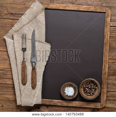 View from above of spices vintage spoon and fork on linen napkin on a blackboard on a rustic wooden table with copy space.