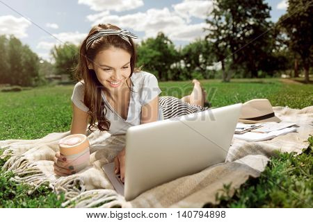 On the emotional wave. Cheerful charming delighted woman smiling and using laptop while resting on the grass