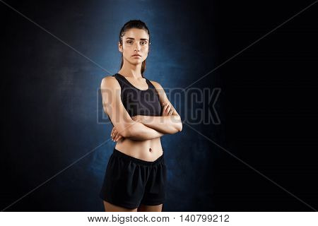 Young beautiful sportive girl posing with crossed arms over dark background. Copy space.