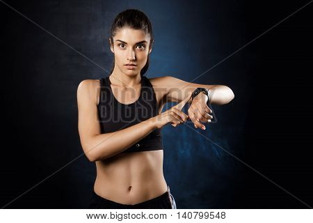 Young beautiful brunette sportive girl pointing at watch over dark background. Copy space.