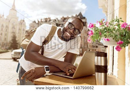 Positive thinker. Cheerful handsome smiling man leaning on the table and using laptop while standing outside the cafe