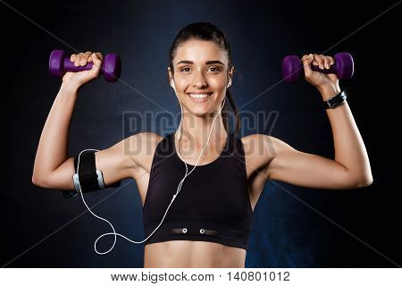 Young beautiful bruntete sportive girl training with dumbbells over dark background. Copy space.