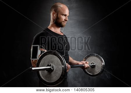 Young powerful sportsman in heaphones training with barbell over black background. Copy space.