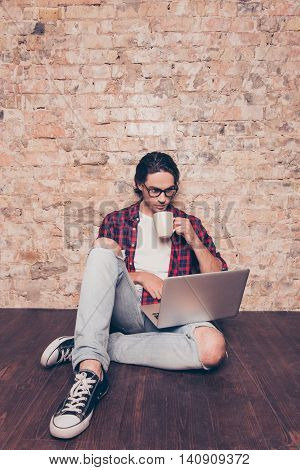 Handsome Young Man In Glasses Sitting On Floor With Laptop And Cup Of Coffee