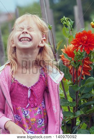 a little girl of preschool age in a pink shirt ,stand near a red chrysanthemum flower in the garden, long blond hair, portrait,laugh, nicker,