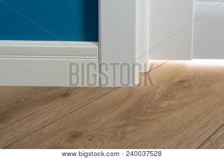 Details In The Interior. Laminated Parquet Floors Immitating Oak Texture, White Baseboard, White Doo