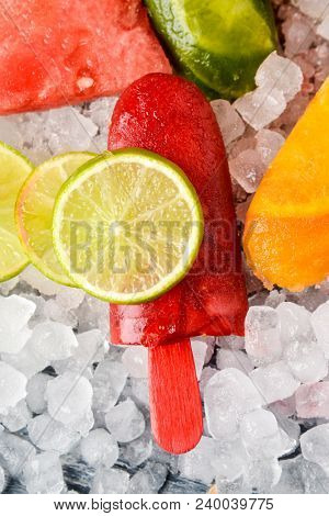 high angle view of some different homemade ice pops, made with different natural fruit juices and pieces of fruit, such as watermelon, lime or orange, placed on crushed ice