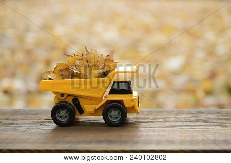 A Small Toy Yellow Truck Is Loaded With Yellow Fallen Leaves. The Car Stands On A Wooden Surface Aga