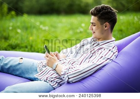 Handsome Young Smile Man Lying On Inflatable Sofa Lamzac Typing On Phone While Resting On Grass In P