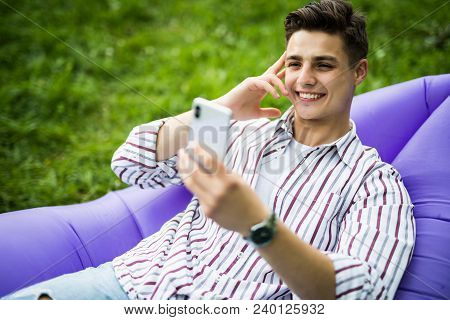 Handsome Young Man Lying On Inflatable Sofa Lamzac Read News On Phone While Resting On Grass In Park