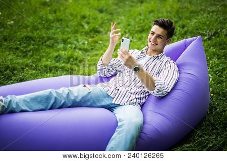 Handsome Young Man Lying On Inflatable Sofa Lamzac Use On Phone While Resting On Grass In Park