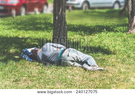 Old Barefoot Homeless Or Refugee Man Sleeping On The Grass In The City Park Using His Travel Bag As 