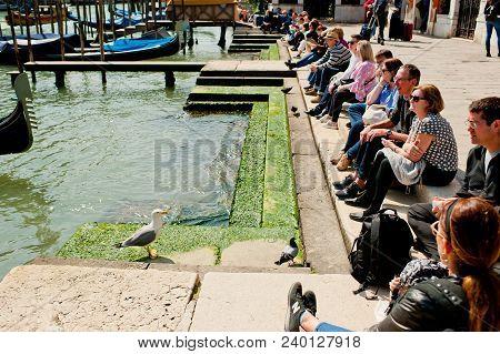 Venice, Italy - April 06, 2018: Tourists Sit On The Bank Of The Canal.