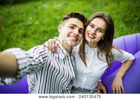 Young Couple Sitting On Inflatable Sofa Lamzac And Take Selfie On Phone While Resting On Grass In Pa