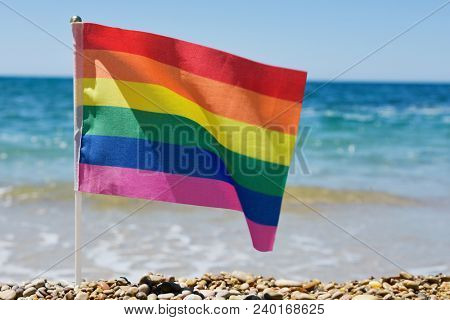 closeup of a small rainbow flag in a pole stuck in a shingle beach, with the ocean in the background