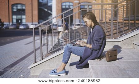 A Young Girl In Jeans In Sunglasses Is Working With A Laptop Outisde Drinking Coffee