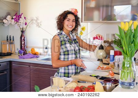Portrait Of Brunette Attractive Female With Curly Hair In A Home Kitchen With A Lot Of Flowers.