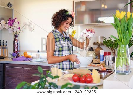 Positive Brunette Female Makes Dough In A Kitchen With Yellow Tulips On Background.