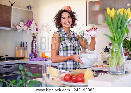 Positive Brunette Female Makes Dough In A Kitchen With Yellow Tulips On Background.