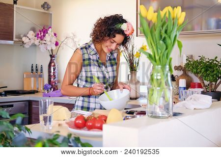 Positive Brunette Female Makes Dough In A Kitchen With Yellow Tulips On Background.