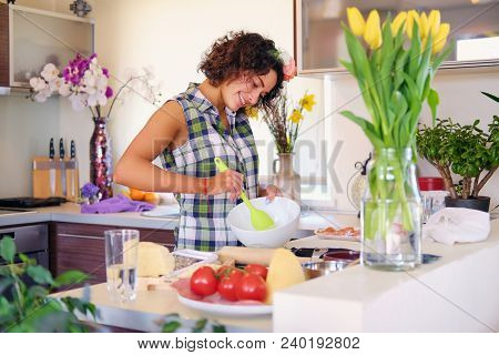 Positive Brunette Female Makes Dough In A Kitchen With Yellow Tulips On Background.