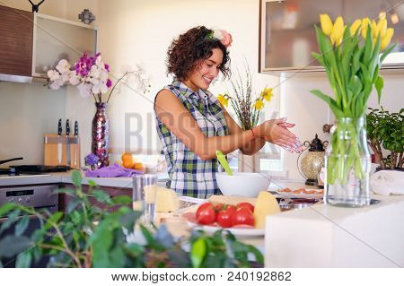 Positive Brunette Female Makes Dough In A Kitchen With Yellow Tulips On Background.