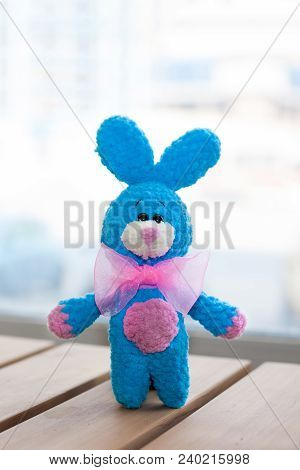 A Small Knitted Blue Rabbit With Pink Bowtie On Wooden Surface. Knitted Toy, Handmade. Amigurumi.