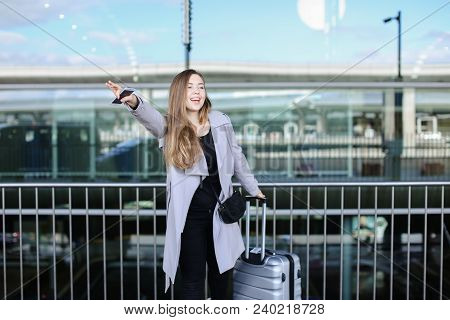 Young Woman Stopping Taxi With Raised Hand, Valise And Smartpone Near Airport. Concept Of Business T
