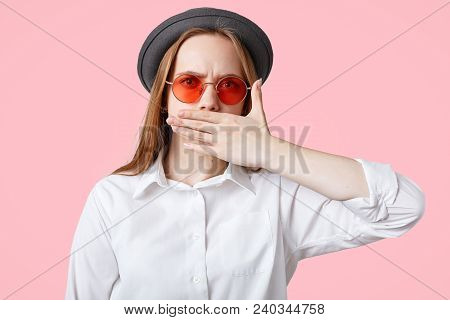 Speachless And Angry Female Covers Mouth As Tries Keep Silence, Wears Elegant Hat And Stylish Shades