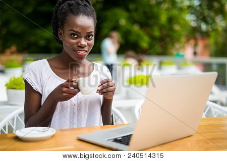 Portrait Of African American Girl In Glasses Sitting At Cafe With Laptop And Holding Cup Of Coffee I